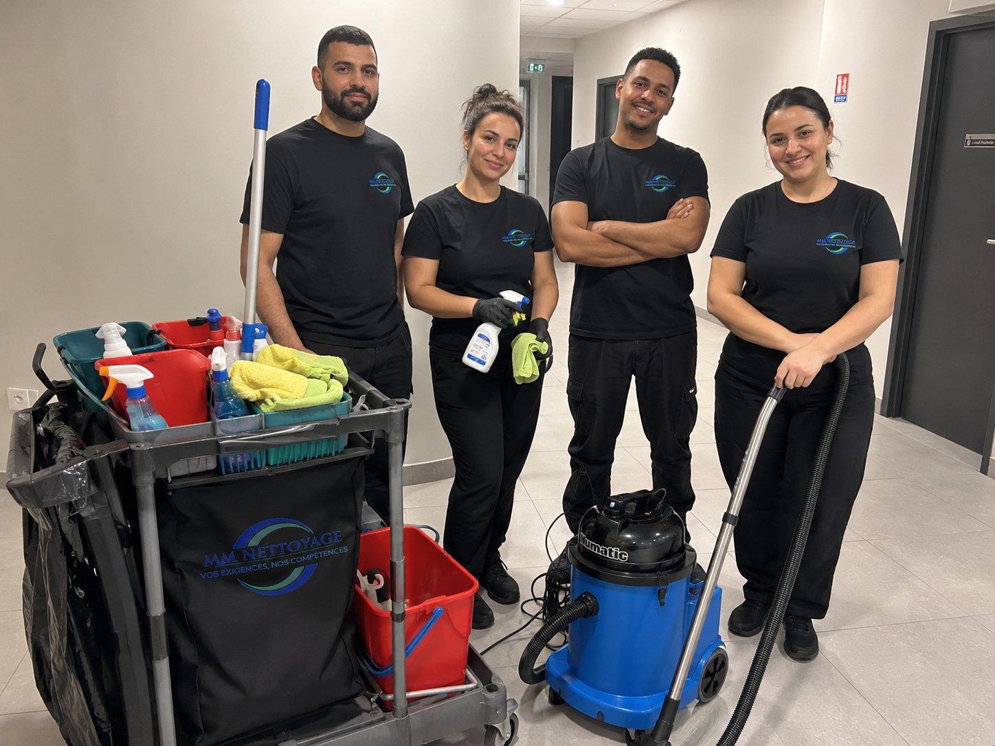 L'équipe MM NETTOYAGE en intervention dans un couloir — t-shirts noirs, chariot de service, aspirateur Numatic
